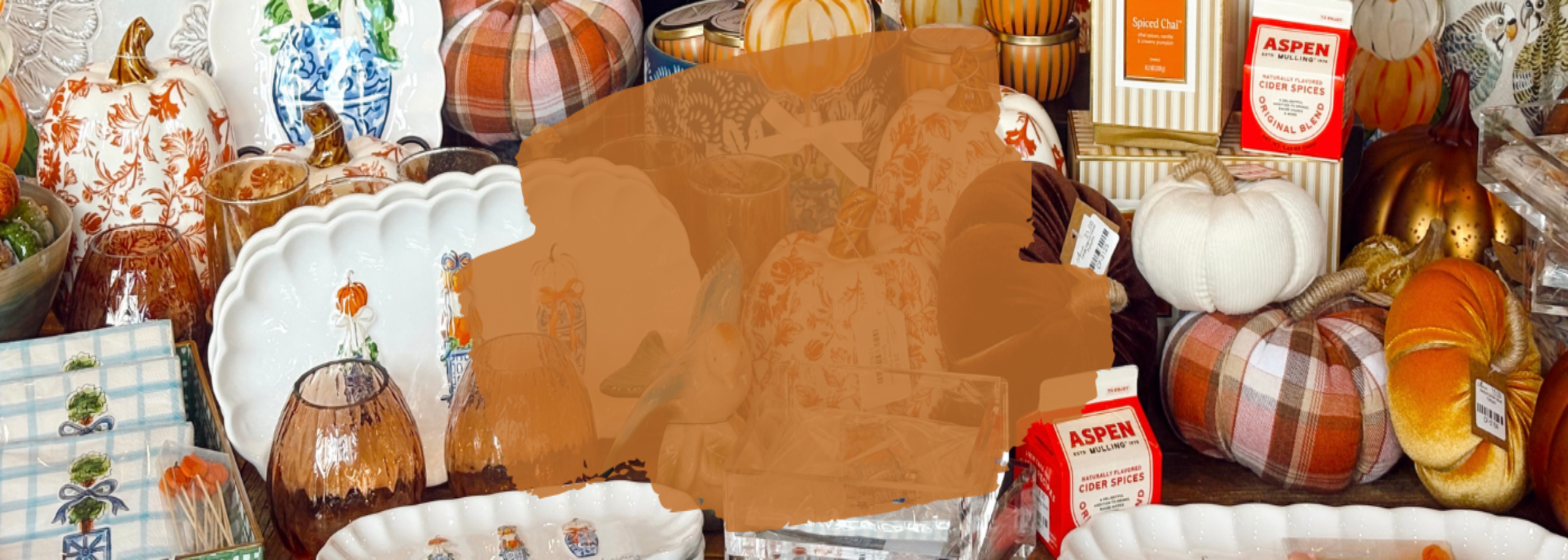 A busy fall-themed tabletop display with pumpkins, plaid fabric, white ceramic dishes, glassware, and small decorative items. A large brown rectangular overlay obscures the center of the image, and several product details are partially hidden.